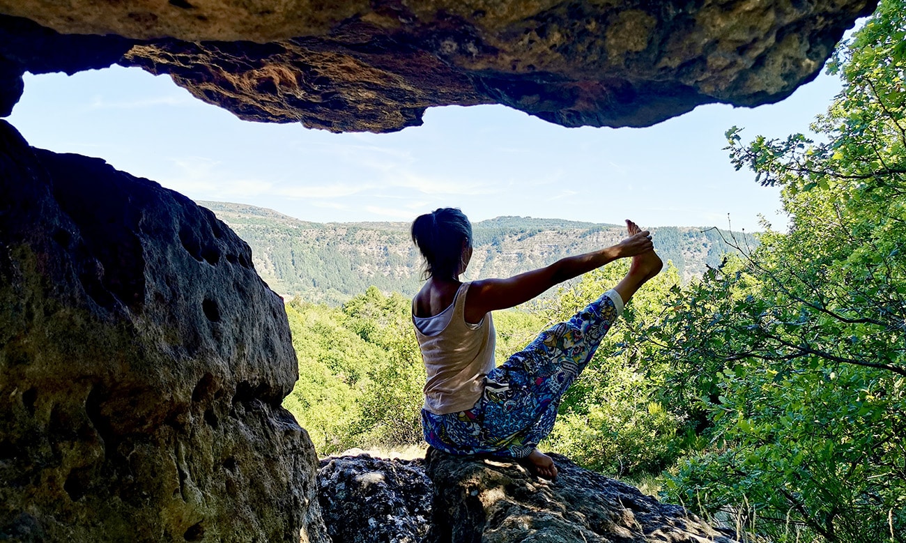 Scylia assise sur un rocher le pied droit dans la main droite en train de faire une pose de yoga du cachemire, au milieu de la nature.