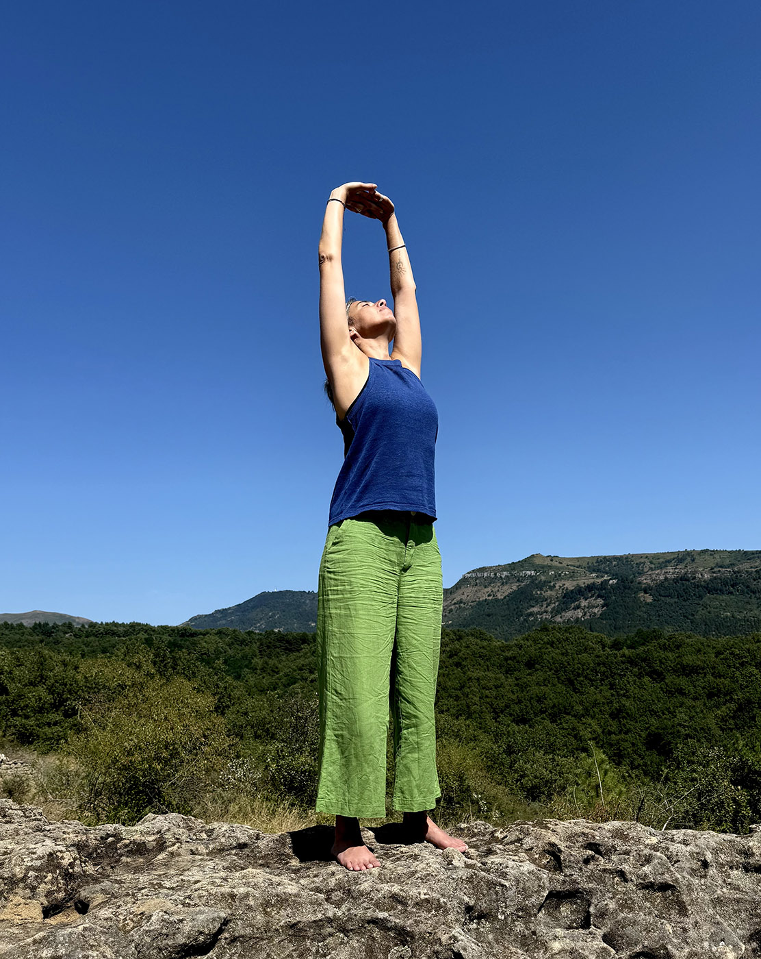 Scylia assise sur un rocher le pied droit dans la main droite en train de faire une pose de yoga du cachemire, au milieu de la nature.